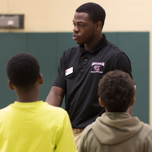 A black male physical education teacher is standing in a gym in front of two elementary school-aged students.