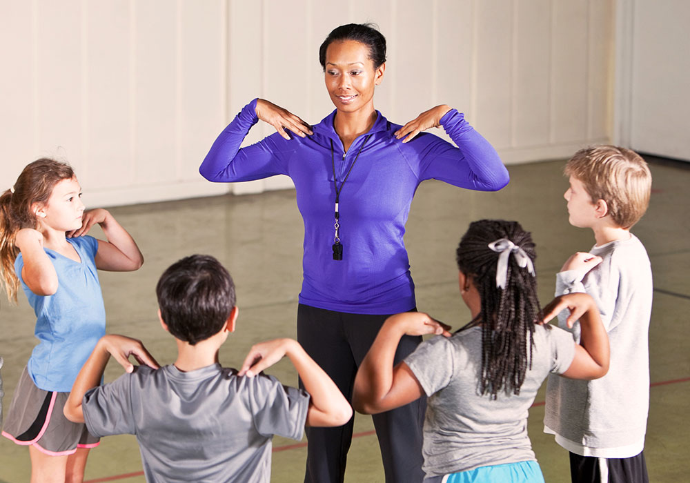 A black female physical education teacher stands at the front of a classroom with her hands on her shoulders. Two elementary school-age students are facing her with their hands on their shoulders.