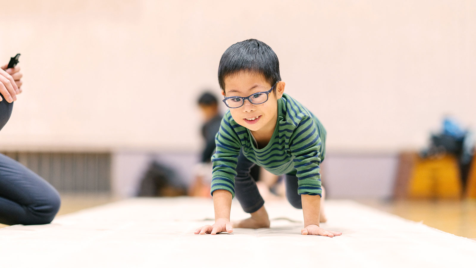 An elementary school-aged boy with dark hair and glasses is crawling toward the camera on a white gym mat.