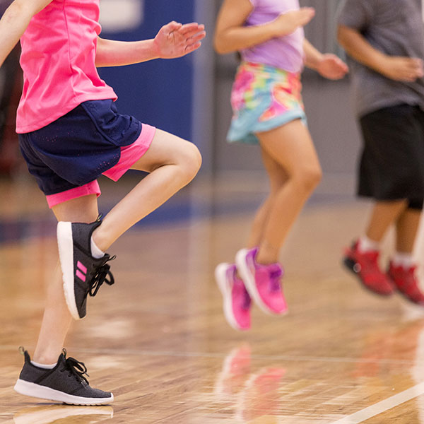 Three children dressed in shorts and tennis shoes stand in a row, jumping on a gym floor.