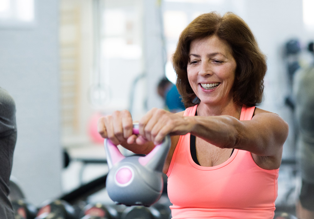 Smiling woman in an exercise facility lifting a kettlebell, illustrating physical therapy used in neurological rehabilitation.