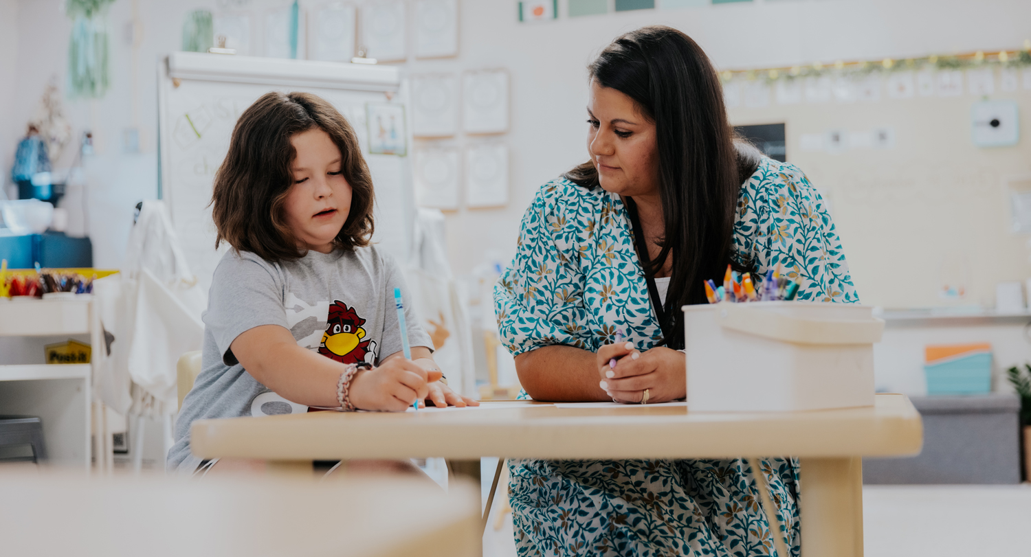A teacher sits at a table with a young student in a classroom.
