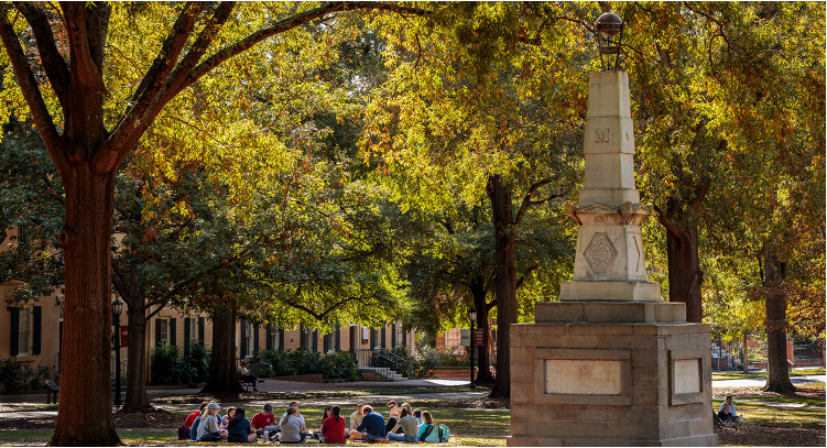 Students gather on the historic Horseshoe at the University of South Carolina, sitting in a circle beneath shaded trees near a stone monument, with campus buildings in the background.
