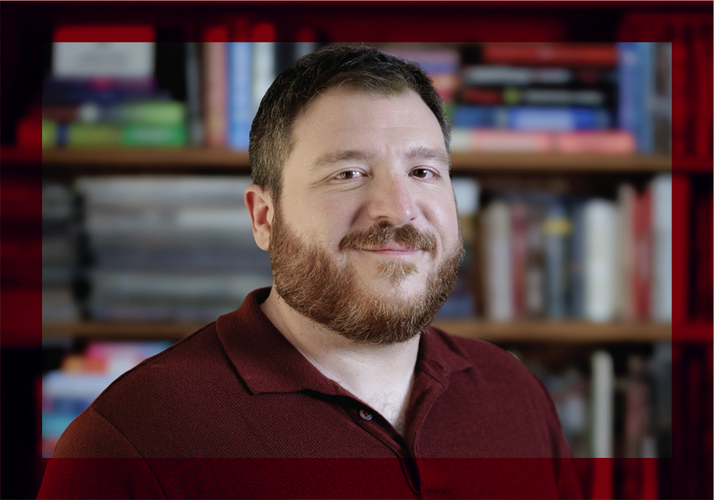 Portrait of a person with short dark hair and a beard wearing a maroon polo shirt, standing in front of a blurred bookshelf background.