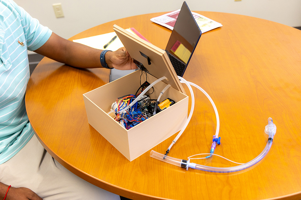 A person holds open a small box that appears to be full of electronic, including sensors and motors. The device is attached to a clear medical tube on the table.