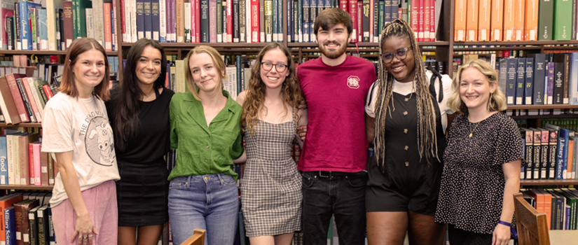 A group of students pose for a photo in front of shelves of books at the library.