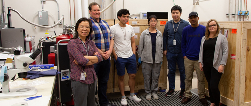 Rongyin Jin and a group of students pose for a photo in a laboratory full of equipment, some of it still packed in boxes taller than an average person.