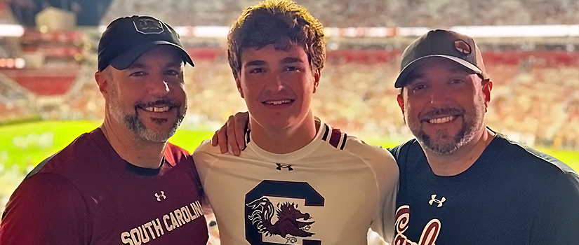 Three smiling men in South Carolina Gamecocks shirts pose together at a night football game, with the brightly lit stadium and field full of fans in the background.