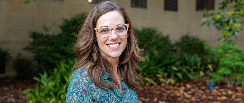 A woman wearing glasses smiles outdoors in front of campus landscaping and a building.
