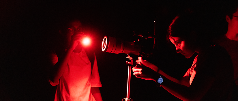 Student adjusts a telescope at night while another uses a red flashlight to preserve night vision during a stargazing event.