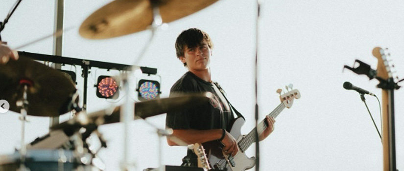 Musician playing bass guitar on stage during a live outdoor performance, with drums and microphone stand in the background