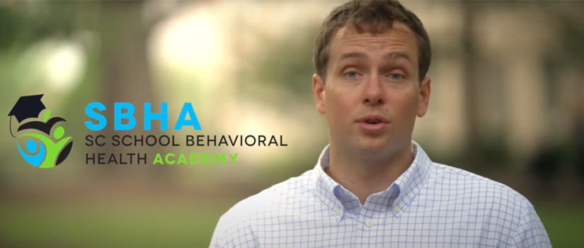 A man in a light blue checkered shirt speaks in front of a blurred outdoor background. The logo for the SC School Behavioral Health Academy (SBHA) is displayed to his left, featuring an icon of a figure in green and blue with a graduation cap. The text reads 'SBHA SC School Behavioral Health Academy' in blue and green.