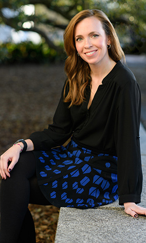 Kathleen Searles, a political communications expert, sits outdoors on a stone bench, smiling warmly. She wears a black blouse and a blue patterned skirt, with a background of greenery and trees in soft focus.