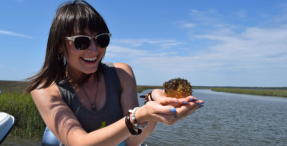 A woman wearing sunglasses and a tank top smiles at a small puffer fish in her hand. Behind her is a river and grassland.