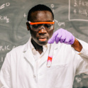 Kwame wears a lab coat and googles and holds a test tube. He is standing in front of a chalk board.