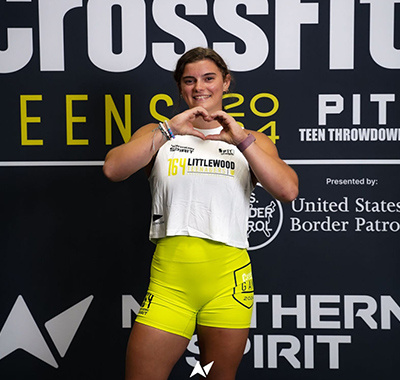 Reese Littlewood, wearing a white tank top and bright yellow shorts, poses confidently in front of a CrossFit Teens 2024 event backdrop, forming a heart shape with her hands