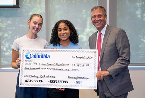 Ella Michel and Tina-Maria Sandoval holding a large check for $4,779 awarded by the City of Columbia, presented by Columbia Mayor Daniel Rickenmann. They are standing indoors with a backdrop of windows showing greenery outside.