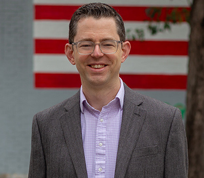 photo of Bryan Levy standing outside among a metal sculpture with an american flag behind him. 