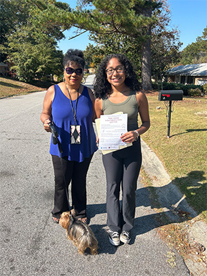 Tina-Maria Sandoval and her mother standing on a residential street holding flyers about weatherization services. Tina-Maria's mother holds a small dog on a leash, and houses with trees are visible in the background.