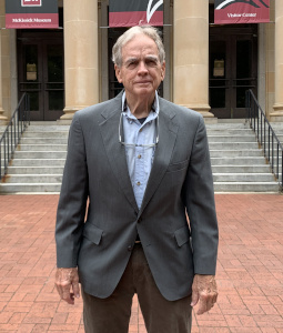 Don Jordan wears a gray blazer and blue button down shirt with his glasses around his neck. He stands with his arms to the side in front of McKissick Museum's steps and columns.