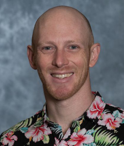 John wears a black and pink Hawaiian print shirt and smiles at the camera