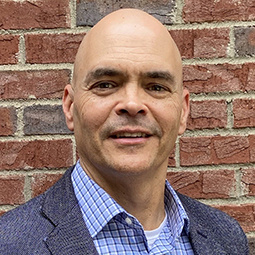 a man smiling in front a brick wall with a blue blazer on a checked button up shirt