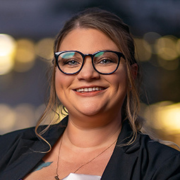 a woman with dark rimmed glasses and a black jacket on smiling with a city background