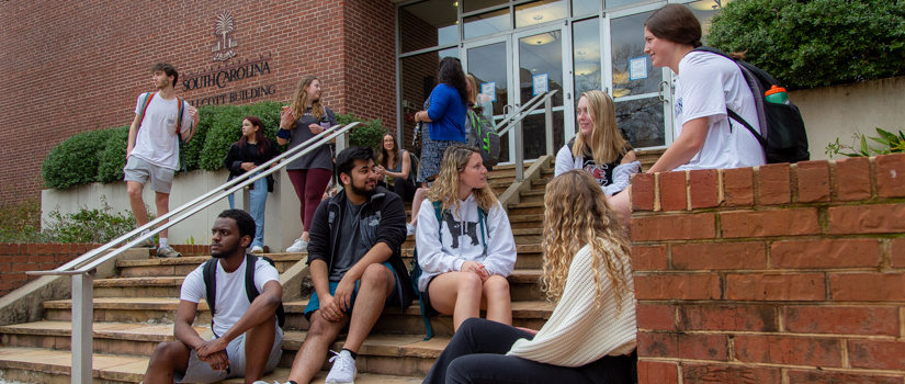 A diverse group of students sit outside on the steps in front of a brick building. They are split off in various smaller groups and are engaged in conversation with each other.