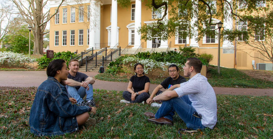 Five students sit in a circle on the grass in front of Barnwell College, smiling and chatting with each other.
