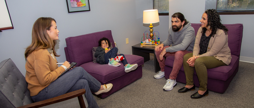 A clinician sits in a private room with a family of three. They are sitting on comfortable furniture while the child plays and the adults talk.