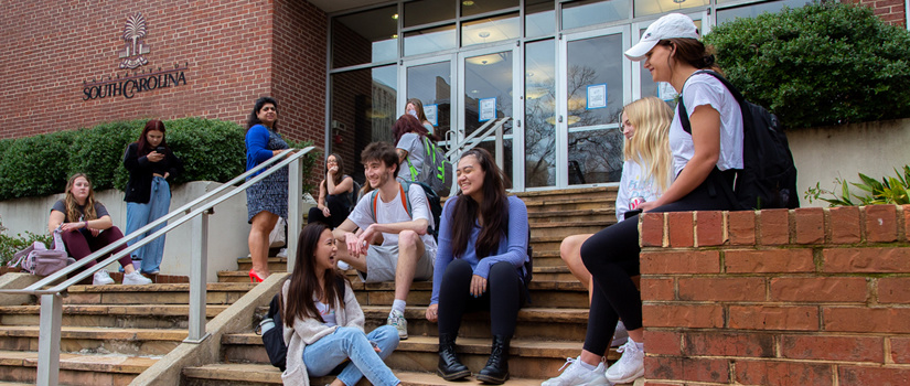 A wide shot photograph shows a diverse group of students are gathered on the front steps of a brick building. Some are standing and some are sitting together as they chat and laugh.