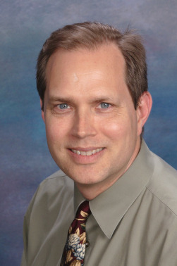 Jeffrey Schatz wears a gray shirt with a multi-color tie and smiles at the camera against a blue background.