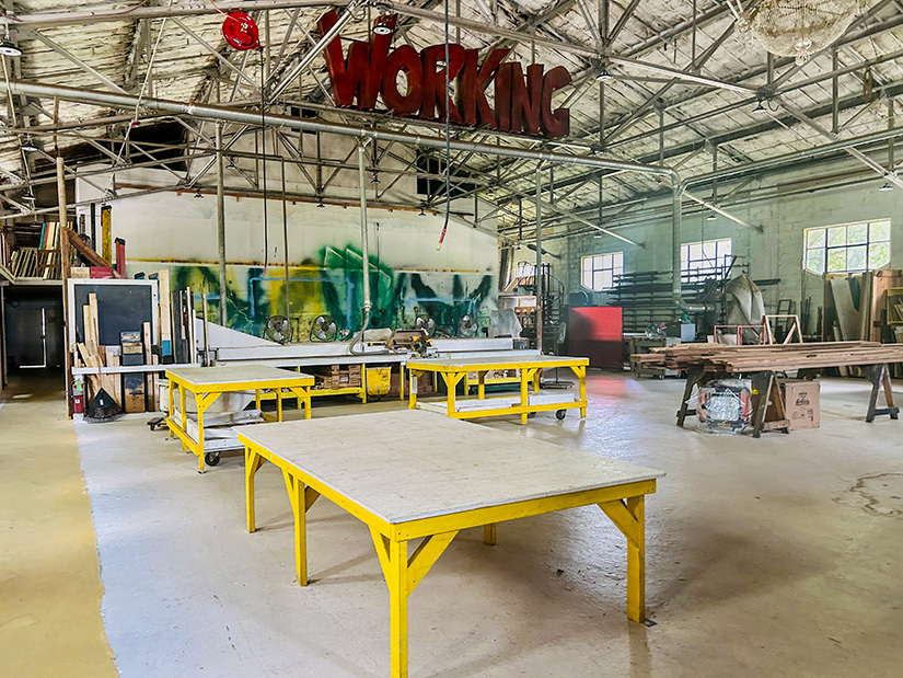 Scenic shop at USC. Work table in the foreground with additional tables in the background. A sculpture of the word Working is hung from the ceiling.