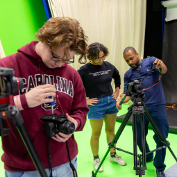 A student works on a camera; a student and professor look at a camera in the background.
