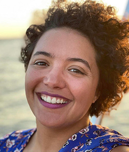 a photo of a woman with dark curly hair smiling in front of a body of water