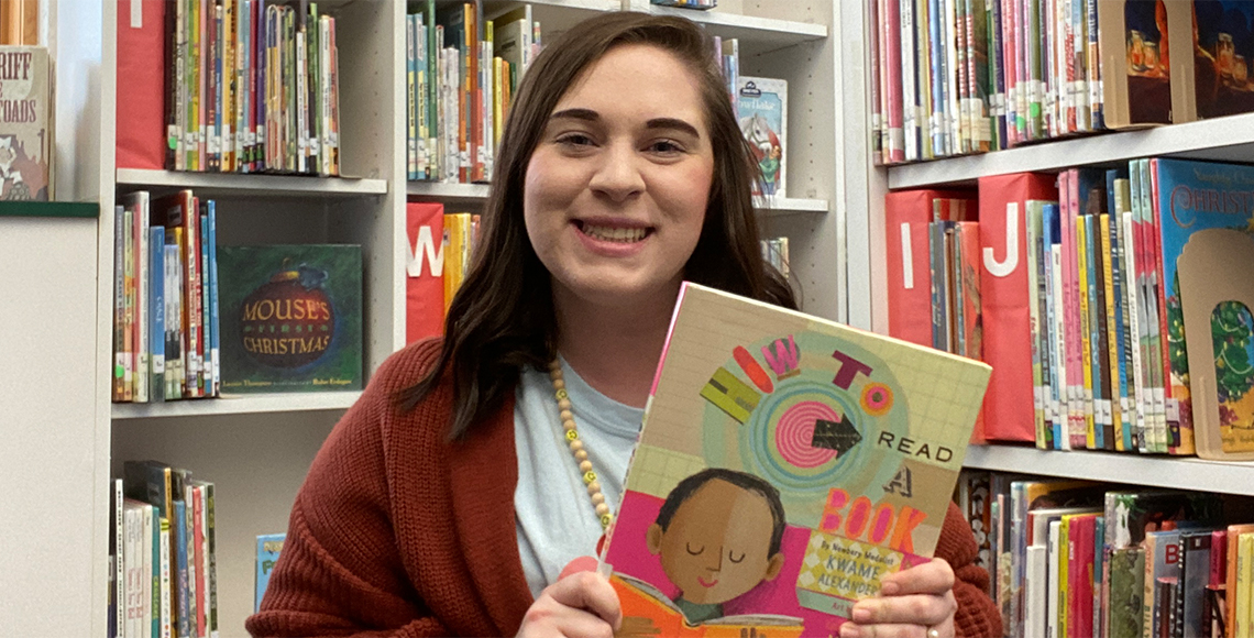 Megan Evans holding a book while sitting on the floor in a library.