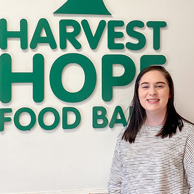 Megan Evans standing in front of a sign which reads harvest Hope Food Bank