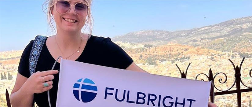 Blonde woman in front of a wrought-iron fence, holding a sign that says "Fulbright"