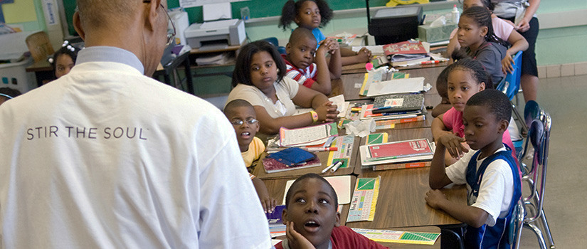 An African American teacher wearing a shirt that reads 'Stir the Soul' on the back stands in front of a classroom full of children.
