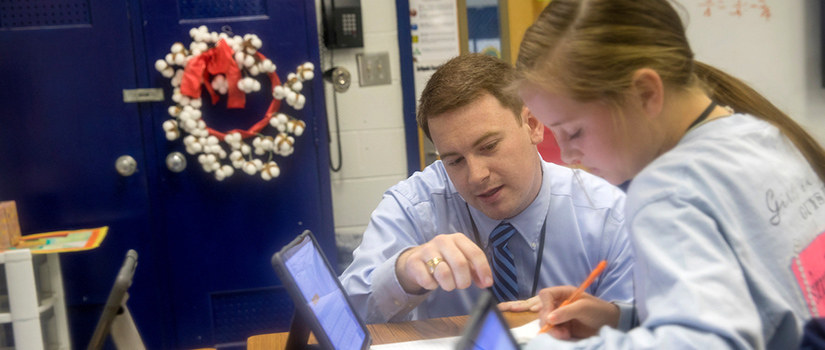 a white male teacher instructs a white female middle school student working on a tablet.