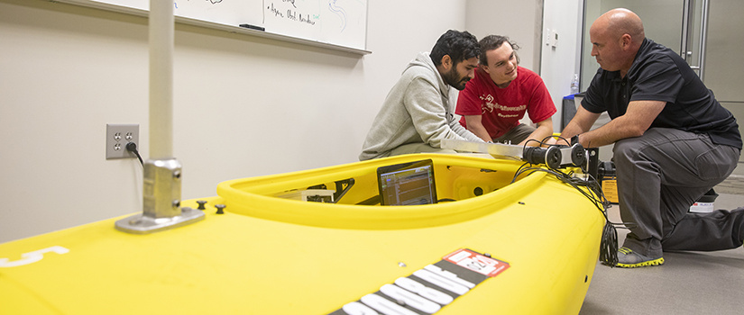 students and professor kneel next to robotic kayak