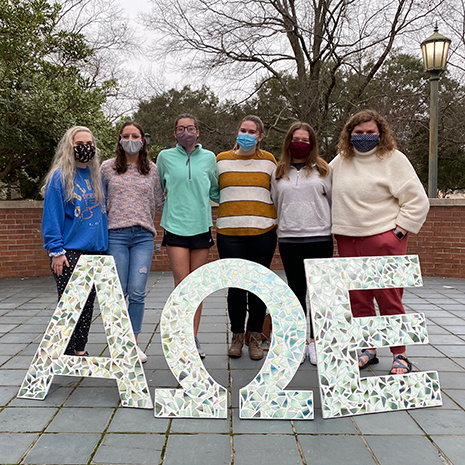 six women stand outdoors wearing masks behind large AEO letters