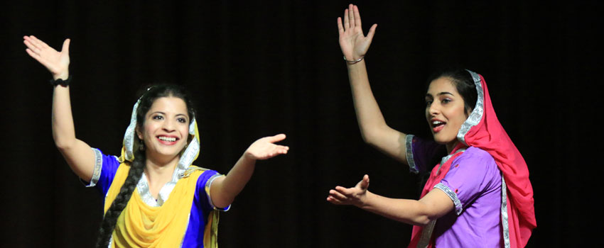 Two women in colorful traditional attire perform a dance on stage. The woman on the left wears a yellow and blue outfit with a white headscarf, while the woman on the right wears a purple outfit with a pink headscarf. Both have their arms raised and are smiling joyfully against a dark background.
