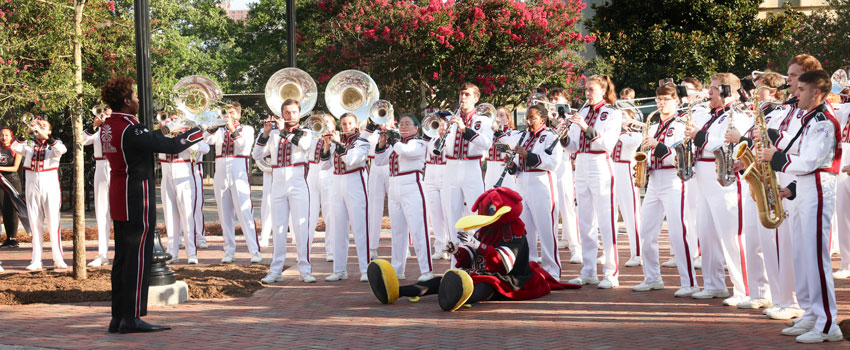 The USC marching band dressed in white uniforms with garnet and black accents performs outdoors on the historic Horseshoe. In front of the band, Cocky is sitting on the ground, engaging with the audience.