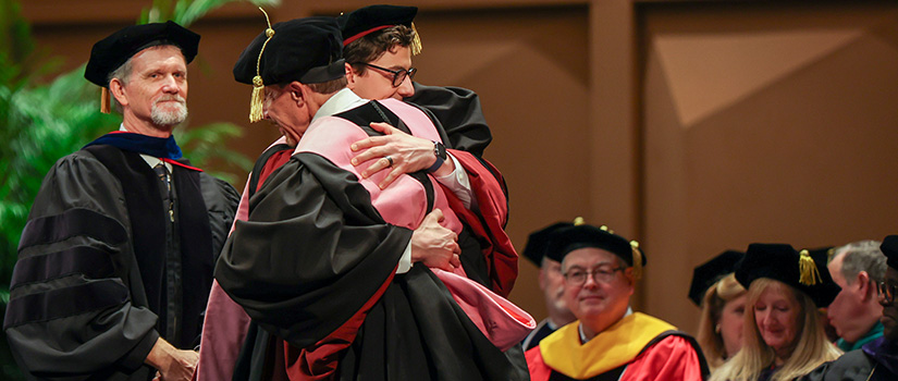 Graduate embraces advisor on stage during doctoral hooding ceremony