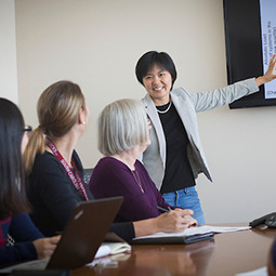 A female Asian presenter disucsses material on a projector with three other women one of whom is wearing an official UofSC lanyard and one of whom is also of Asian descent.