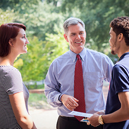 Two students one male and one female talking to a male professor outside with greenery in the background.