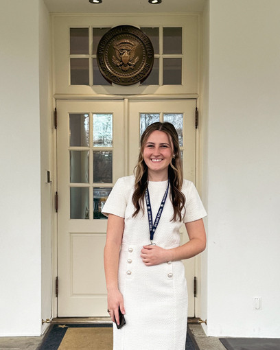 Blair Elliott smiling in front of the West Wing door in Washington D.C. The Seal of the President of the United States is seen in the background.