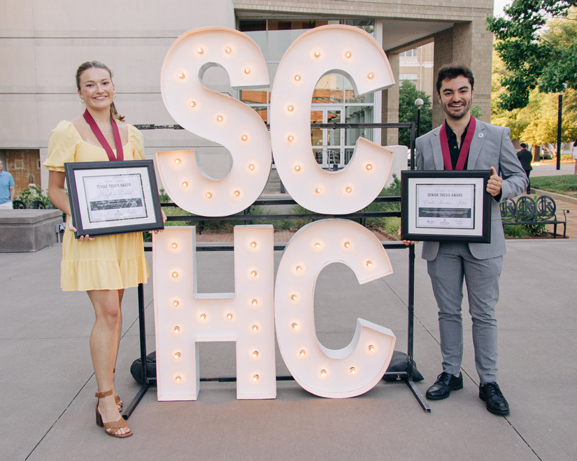 Meaghan Arnold and Carlos Sanchez-Julia posing next to large, lit up 'SCHC' block letters as they hold their award certificates.
