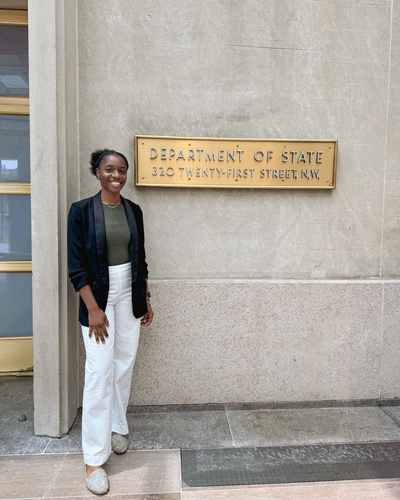 woman standing in front of concrete wall with gold plaque next to her that reads "Department of State"